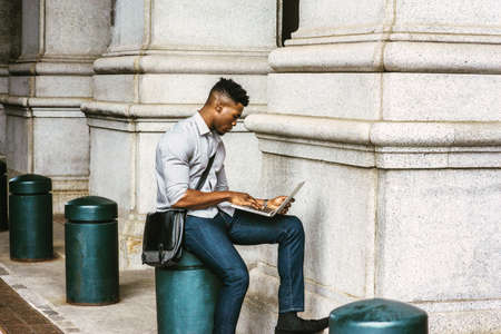 African American College Student studying in New York, wearing shirt, jeans, carrying shoulder leather bag, sitting on pillar on street, reading, working on laptop computer.  filtered effect.の写真素材