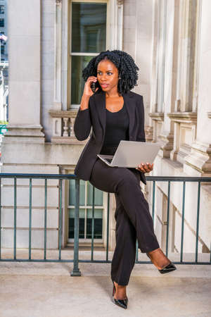 African American Businesswoman working in New York. Young black college student with braid hairstyle sitting on railing in vintage style office building, working on laptop computer, making phone call.の写真素材