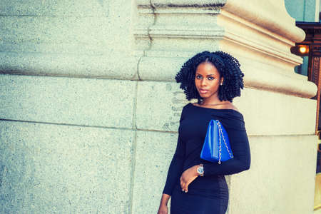 African American Businesswoman working in New York. Wearing long sleeve, slim off shoulder dress, carrying blue bag under arm, young black lady with braid hairstyle standing on street, looking at you.の写真素材