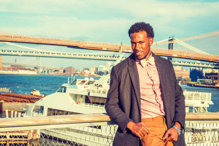 African American Businessman traveling in New York, wearing woolen overcoat, pink shirt, brown pants, bow tie, standing at harbor, looking at you. Manhattan, Brooklyn bridges, boat on backgroundの写真素材