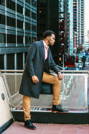 African American Man Winter Fashion in New York, wearing woolen overcoat, pink undershirt, brown pants, leather boot shoes, bow tie, standing by railing on balcony, facing street with high buildings.の写真素材