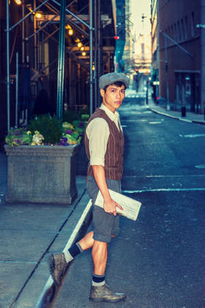 City Boy. Wearing newsboy cap, shirt, patterned vest, gray pants, boot shoes, holding newspaper, Asian American college student walking, crossing narrow street in New York.の写真素材