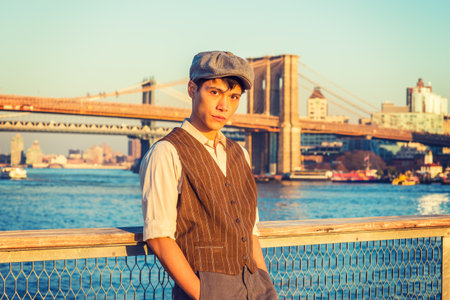 New York City Boy. Wearing newsboy cap, light yellow shirt, patterned vest, Asian American college student standing at harbor in sunset. Manhattan, Brooklyn bridges on background.の写真素材