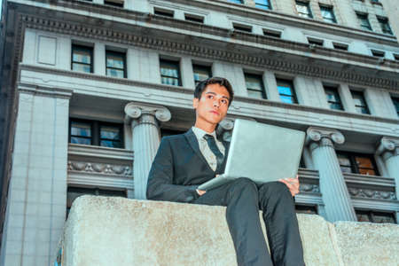 Asian American college student studying in New York. Dressing formally in black suit, necktie, young businessman sitting on street with vintage style building, reading, working on laptop computer,の写真素材