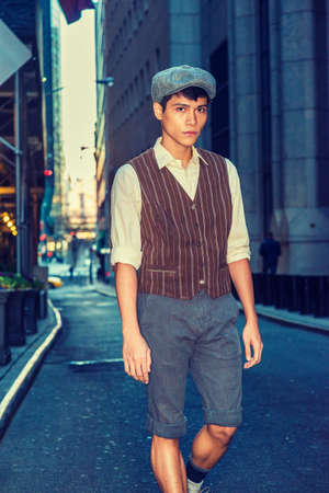 City Boy. Wearing newsboy cap, light yellow shirt, patterned vest, gray pants, Asian American college student walking on narrow street with high buildings in New York.の写真素材