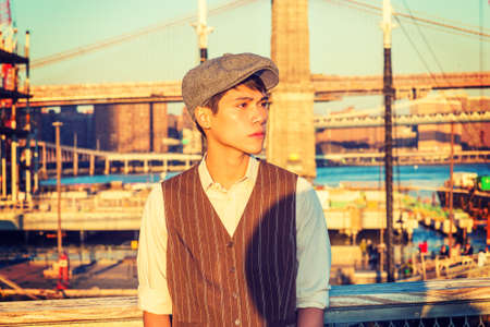 New York City Boy. Wearing newsboy cap, light yellow shirt, striped vest, Asian American college student standing at harbor in sunset, thinking. Bridge, boats on background.の写真素材