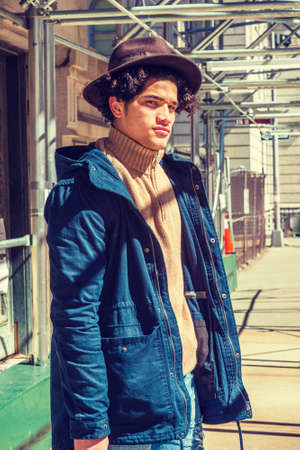 American Man traveling, walking through sidewalk bridge in New York. Wearing blue jacket with hood, knitted turtleneck sweater, Fedora hat, guy with freckle face, curly hair, looking at you under sun.の写真素材