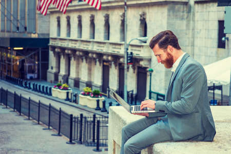 American Businessman with beard, mustache traveling, working in New York, wearing cadet blue suit, sitting outside office building, looking down, working on laptop computer.の写真素材