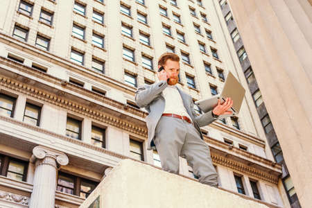 American Businessman with beard, mustache traveling, working in New York, wearing cadet blue suit, standing on street with vintage buildings, working on laptop computer, calling on cell phone.の写真素材