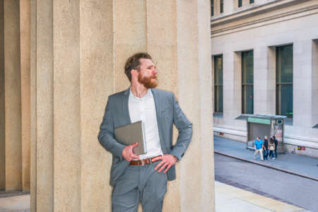 American Businessman with beard, mustache working in New York, wearing cadet blue suit, white undershirt, carrying laptop computer, standing against column outside office building, looking at street.の写真素材