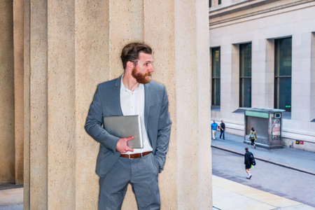 American Businessman with beard, mustache working in New York, wearing cadet blue suit, white undershirt, carrying laptop computer, standing against column outside office building, looking at street.の写真素材