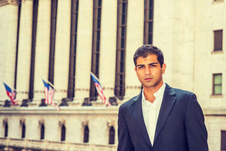 East Indian American Business Man traveling, working in New York. Wearing black suit, a college student standing on street with vintage buildings with flags, looking at you.の写真素材