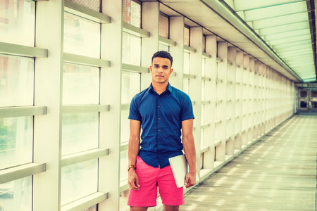 Summer in School. Wearing short sleeve shirt, red shorts, holding laptop computer, African American student standing inside indoor walkway with glass walls, ceiling, wooden floor on campus in New Yorkの写真素材