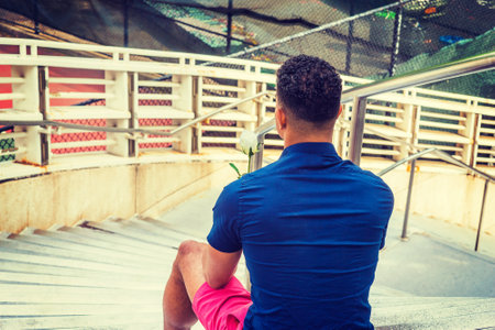 I missing you. Wearing blue short sleeve shirt, red shorts, lonely African American guy sitting on stairs in corner of street, holding white rose, thinking. Back View.の写真素材