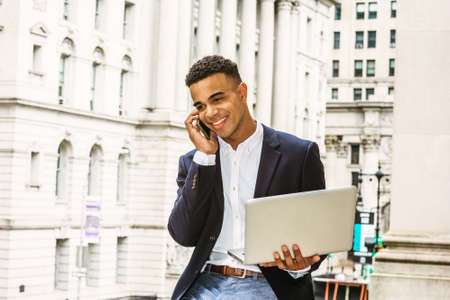 African American Businessman working in New York, wearing black blazer, white shirt, sitting by vintage office building, working on laptop computer, talking on cell phone.の写真素材