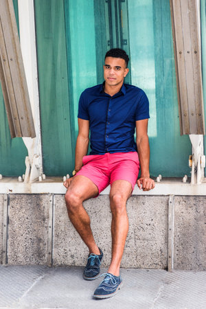 Wearing blue short sleeve shirt, red shorts, sneakers, young African American man sits on street corner by structure in New York in summer, relaxes, takes break. Filtered look with blue tint.の写真素材