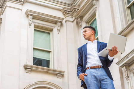 Way to Success. African American male college student studies in New York. Wearing blue blazer, white shirt,  man stands outside vintage office building, works on laptop computer, looks up, thinks.の写真素材