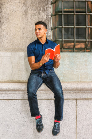 African American College Student studies in New York. Wearing blue short sleeve shirt, jeans, red socks, sneakers, young man sits against vintage wall on street, reads red book.の写真素材