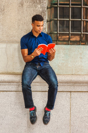 African American College Student studies in New York. Wearing blue short sleeve shirt, jeans, red socks, sneakers, young man sits against vintage wall on street, reads red book.の写真素材