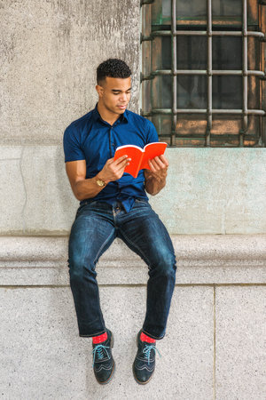 African American College Student studies in New York. Wearing blue short sleeve shirt, jeans, red socks, sneakers, young man sits against vintage wall on street, reads red book.の写真素材