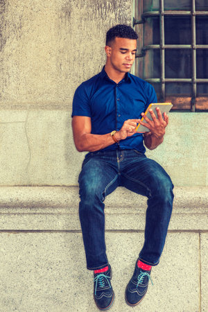 Modern Reading. Wearing blue short sleeve shirt, jeans, red socks, sneakers, African American man sits against vintage wall on street in New York, reads small tablet computer.の写真素材