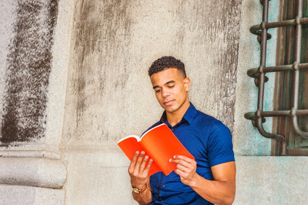 I love reading book. African American College Student studies in New York. Young man wearing blue short sleeve short, sits against vintage wall on campus, looks down, concentratedly reads red book.の写真素材