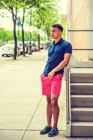 Wearing blue short sleeve shirt, red short, sneakers, young African American man  stands by metal stairs on street in summer, relaxes, thinks. Cars parked on street on background.の写真素材