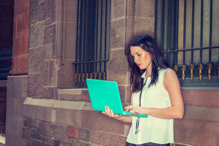 American female college student with long black hair studying in New York in summer, wearing white sleeveless shirt, standing against vintage wall on street, reading, working on laptop computer.の写真素材
