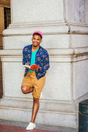 African American college student studying in New York, wearing blue patterned jacket, yellow brown shorts, white sneakers, pink cap, standing against column on campus, reading red book, thinking.の写真素材