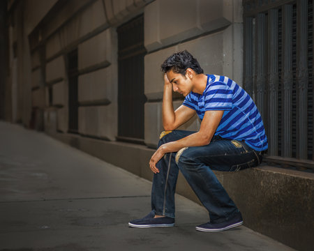 Pensive Young Man in Urban Setting. A young man sits on a stone ledge with a thoughtful expression. The street is deserted, adding to the contemplative atmosphere.の写真素材