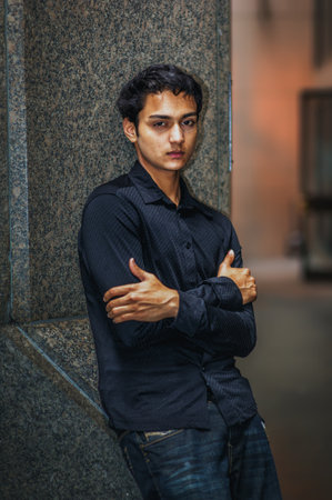 Young Man Leaning Against Wall. A young man in a dark shirt stands confidently against a stone wall, arms crossed. The background has a blurred, warm tone.の写真素材
