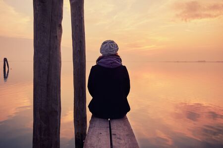 Girl sitting on a pier during a beautiful sunset on the island of Burano near Venice, Italyの写真素材