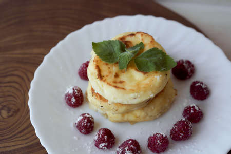 Juicy curd cheesecakes lie on a white saucer with raspberries and mint leaf. Light background. High quality photoの写真素材