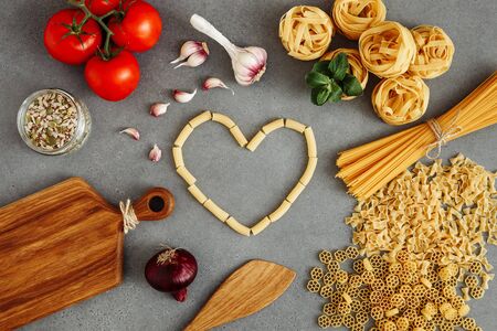 Spaghetti and fettuccine with ingredients for cooking pasta on concrete table with blank of wooden kitchen board, top view. Rustic style. Flat lay. Italian hard pasta heart-shapedの写真素材