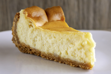 Close up view of a slice of plain cheesecake on a white plate.  Side view.  Wooden background blurred.の写真素材