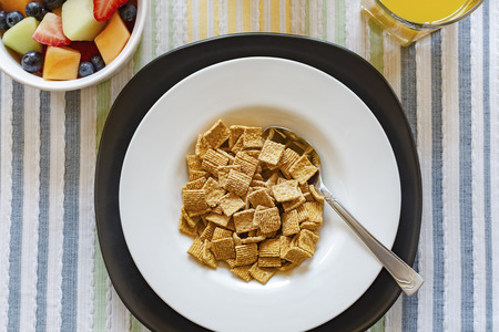 Flat lay of a bowl of cereal with a bowl of mixed fruit in the upper left corner and a glass of orange juice in upper right.  Background of striped multicolored place mat.の写真素材