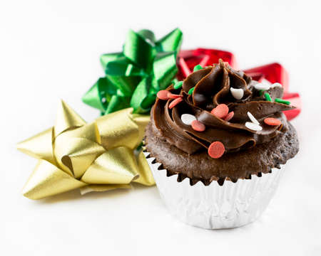 Close up view of a Christmas chocolate cupcake with chocolate frosting and red, green, and white candy sprinkles.  Bows of gold, green and red flank the cupcake.  White background.の写真素材