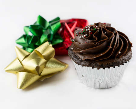 Close up view of a Christmas chocolate cupcake with chocolate frosting and red, green, and white candy sprinkles.  Bows of gold, green and red flank the cupcake.  White background.の写真素材