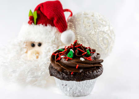 A close up shot of a Christmas chocolate cupcake with candy sprinkles of red, green and white.  Santa and ball ornaments blurred in background.  White background.の写真素材