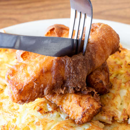 Deep fried fish is being cut into while on a bed of hashbrowns on a white plate.の写真素材