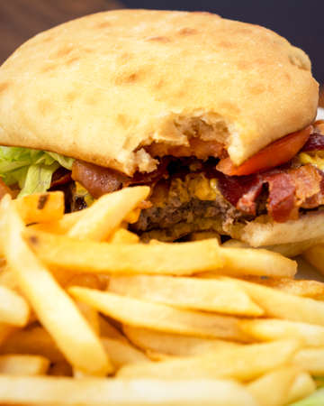 A partron has taken a bite out of a bacon cheeseburger garnished with tomato and lettuce.  French fries are blurred in the foreground.の写真素材