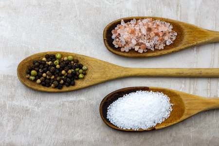Three wooden spoons arranged on a neutral tile.  Spoons filled with pretzel salt, whole peppercorns, and salt.の写真素材