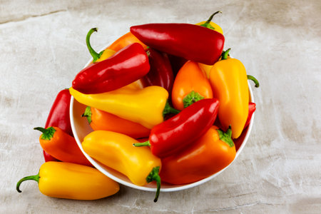 An assortment of red, orange and yellow sweet peppers in a white bowl with a neutral tile background.  Top down view.の写真素材