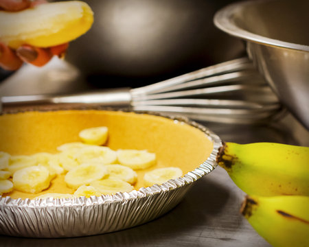 A chef's hand places sliced bananas into a graham cracker pie crust on a stainless steel counter, preparing a fresh banana cream pie in a local cafe.の写真素材