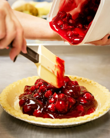 A baker spoons bright red cherry pie filling from a container into a raw, unbaked pastry crust on a stainless steel counter in a cafe kitchen.の写真素材