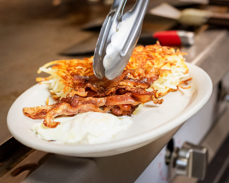 A chef uses tongs to place crispy bacon on a plate of fried eggs and golden hash browns, completing a classic breakfast.の写真素材