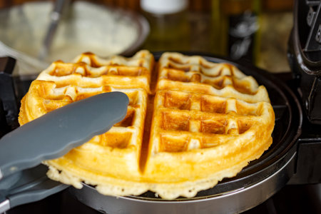 A silicone tong grips a hot, golden-brown waffle being safely removed from the waffle iron, ready to...