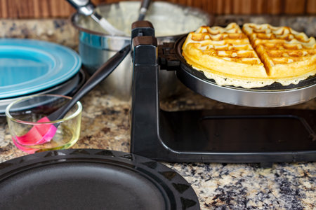 A golden waffle rests on the iron, surrounded by the oil brush, remaining batter, and stacks of...
