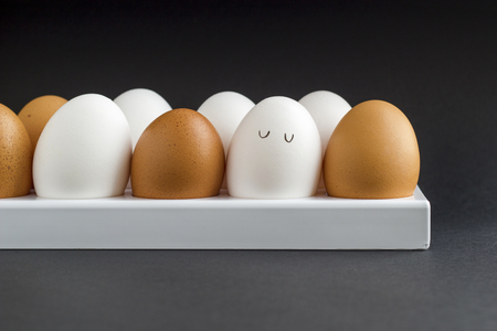 Fresh chicken egg in container on black paper background. Concept for Easter with copy space. White and brown eggs. Painted cute face on a white egg. Drawn cartoon eyes. Frontal view photo.の写真素材