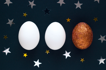 Brilliant and white eggs on dark background with shine lights stars. Top view concept with bronze egg in flat lay style. Idea for greetings and Easter.の写真素材